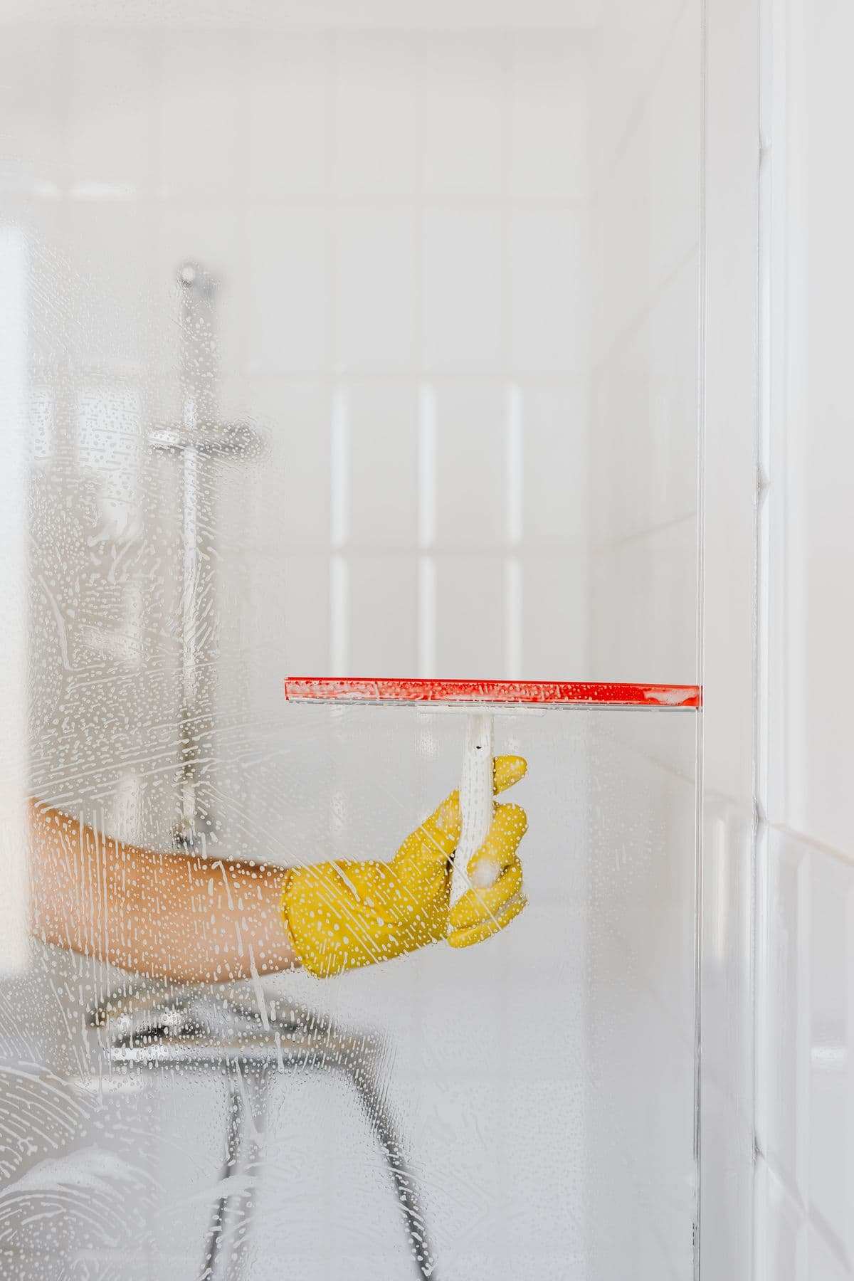 Gloved hand squeegeeing a soap-lathered glass shower screen over white subway tiles