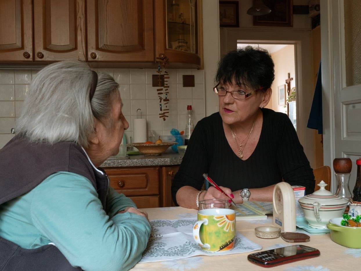 Two older women sitting at a kitchen table sharing tea and conversation in a warm, lived-in home