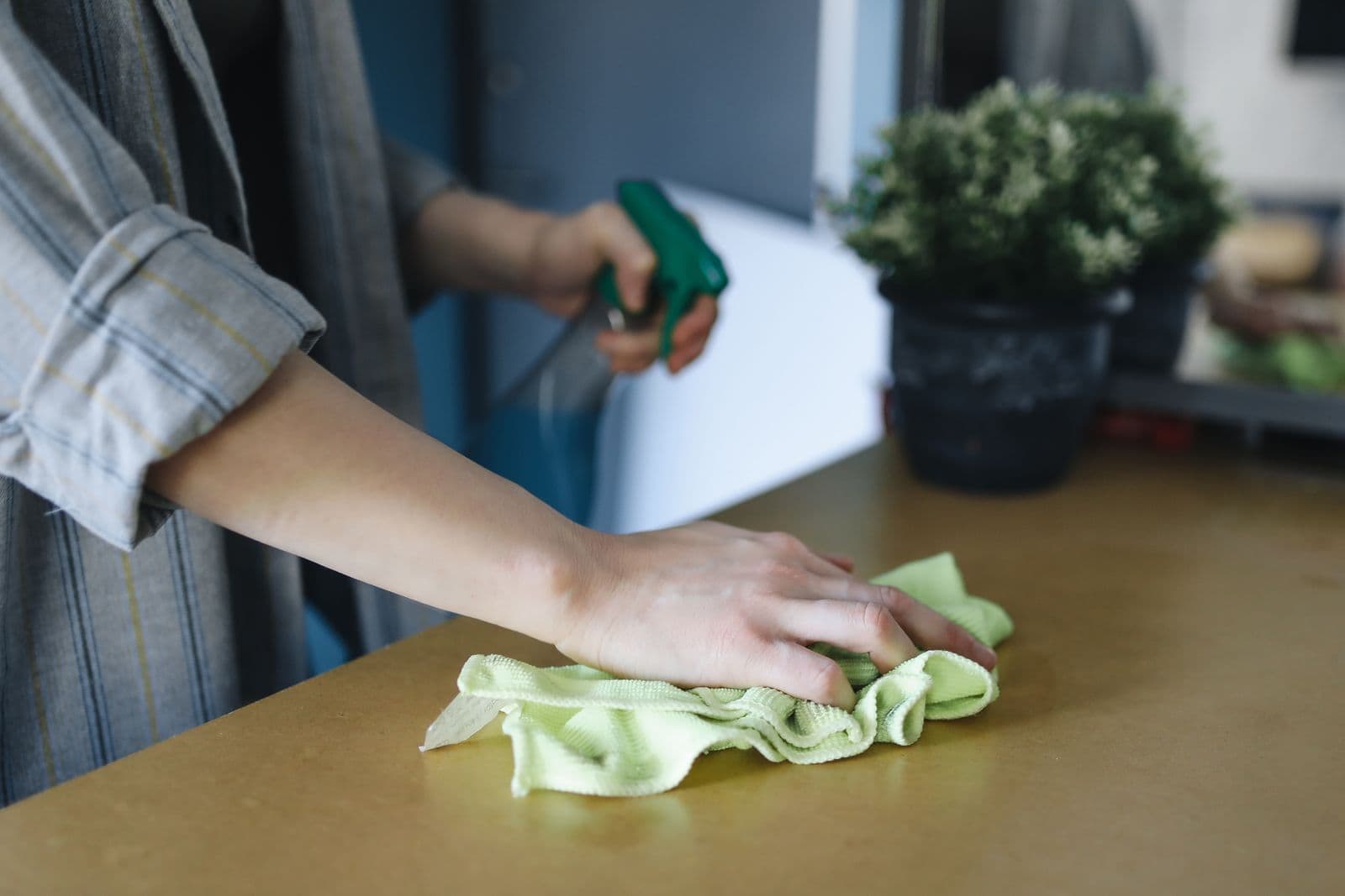 Hand wiping a light wood kitchen countertop with a green microfiber cloth, spray bottle in the other hand, in bright natural light